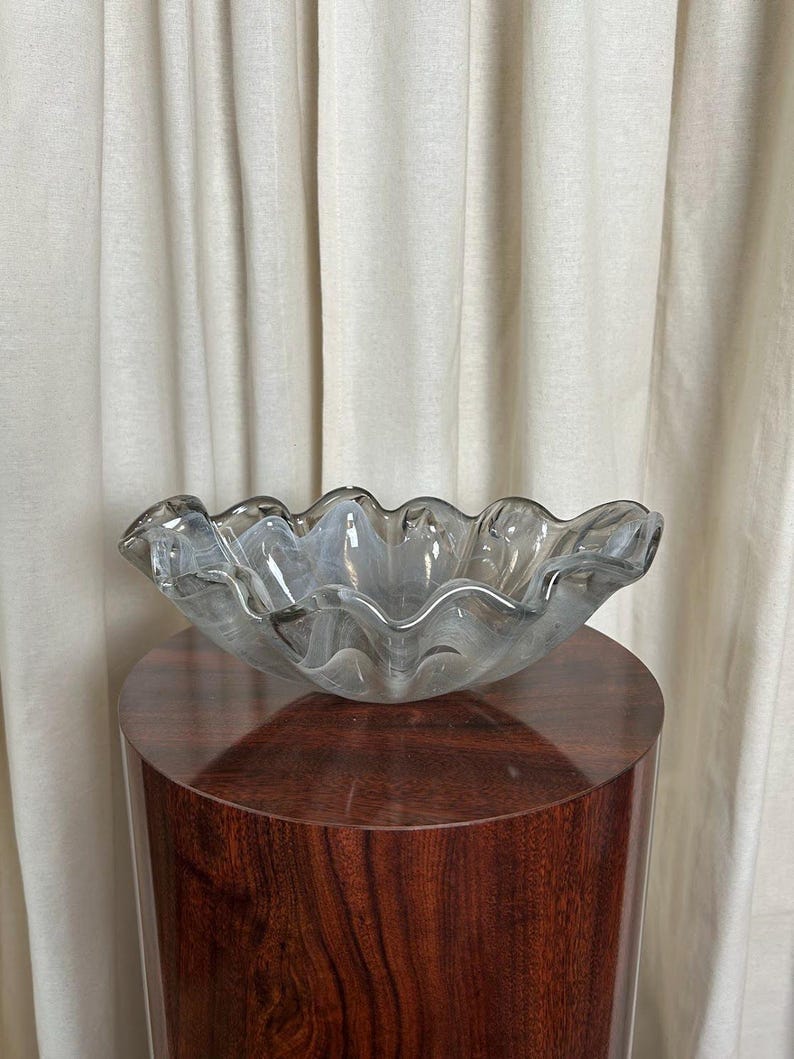 May include: Smoked gray glass bowl with a wavy, scalloped edge. The glass has subtle white swirls throughout. The bowl is displayed on a dark brown pedestal.  The piece is a decorative home accent, ideal for a modern or contemporary interior design style. It is a unique and stylish addition to any living space.