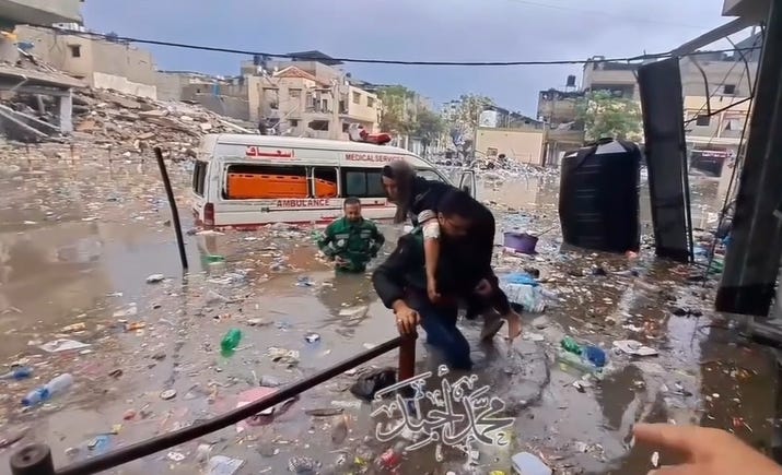 Screen capture of a video. Paramedics evacuate an injured woman who was trapped inside an ambulance in the floods in Jabalia refugee camp, 13.12.23. One of the paramedics helps the woman out of a back window of the ambulance van, and carries her on his back as he wades through water that is almost hip-deep. Rubble is visible in the background and the water is muddy and full of trash and debris.