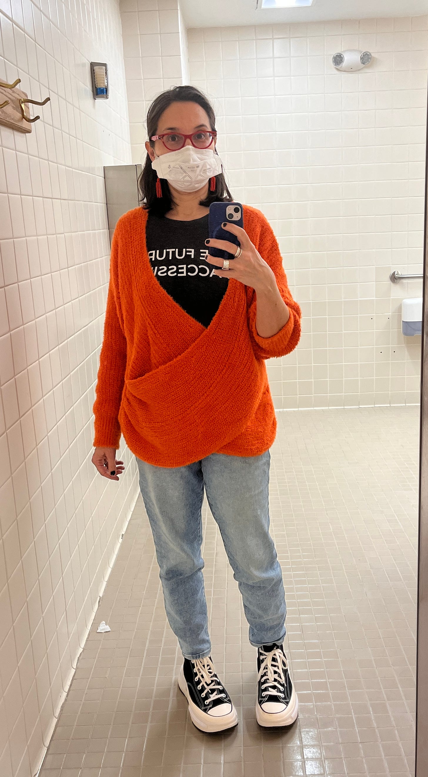 Me, brown shoulder length hair, taking a selfie in a mirror at work, dangly orange earrings, a bright orange sweater, a mask, jeans, and black converse with thick soles. 