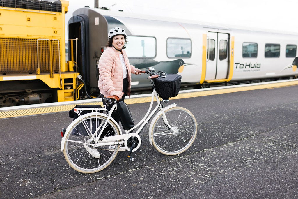 A photograph of Louise at the Frankton train station, standing with their e-bike next to Te Huia. They're looking at the camera and smiling, while the walk their bike alongside the train.
