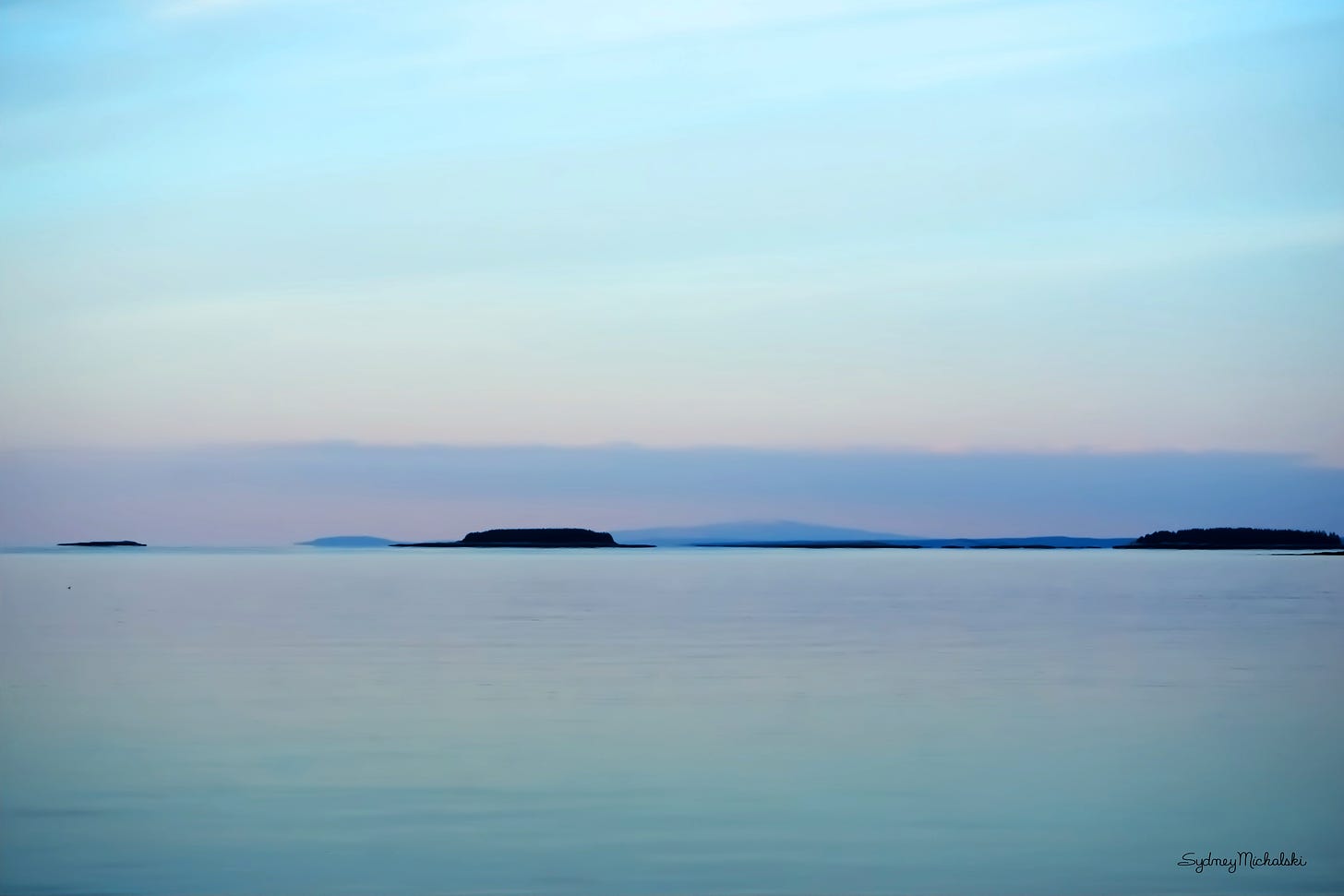 A pastel winter sunrise is reflected in a calm sea with island silhouettes on the horizon.