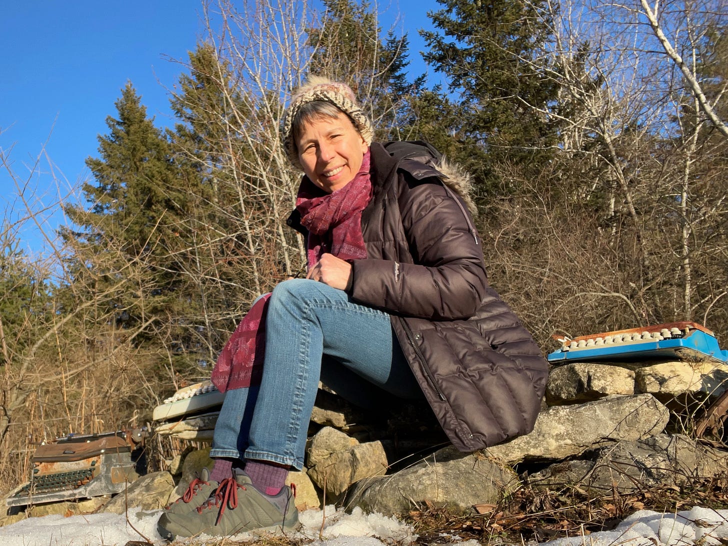 Woman in a jacket and knitted cap sitting outside on some large rocks in the winter with three old rusted typewriters around her, a blue sky overhead and snow in the foreground.