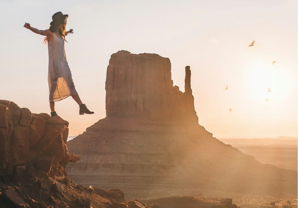 Woman stepping off canyon edge at sunrise with birds flying and large rock formations in the background