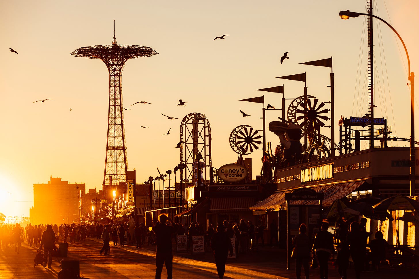 Coney Island at sunset; the image is awash in orange and yellow; people milling about the boardwalk in silhouette; birds flying and the amusements in the near distance.
