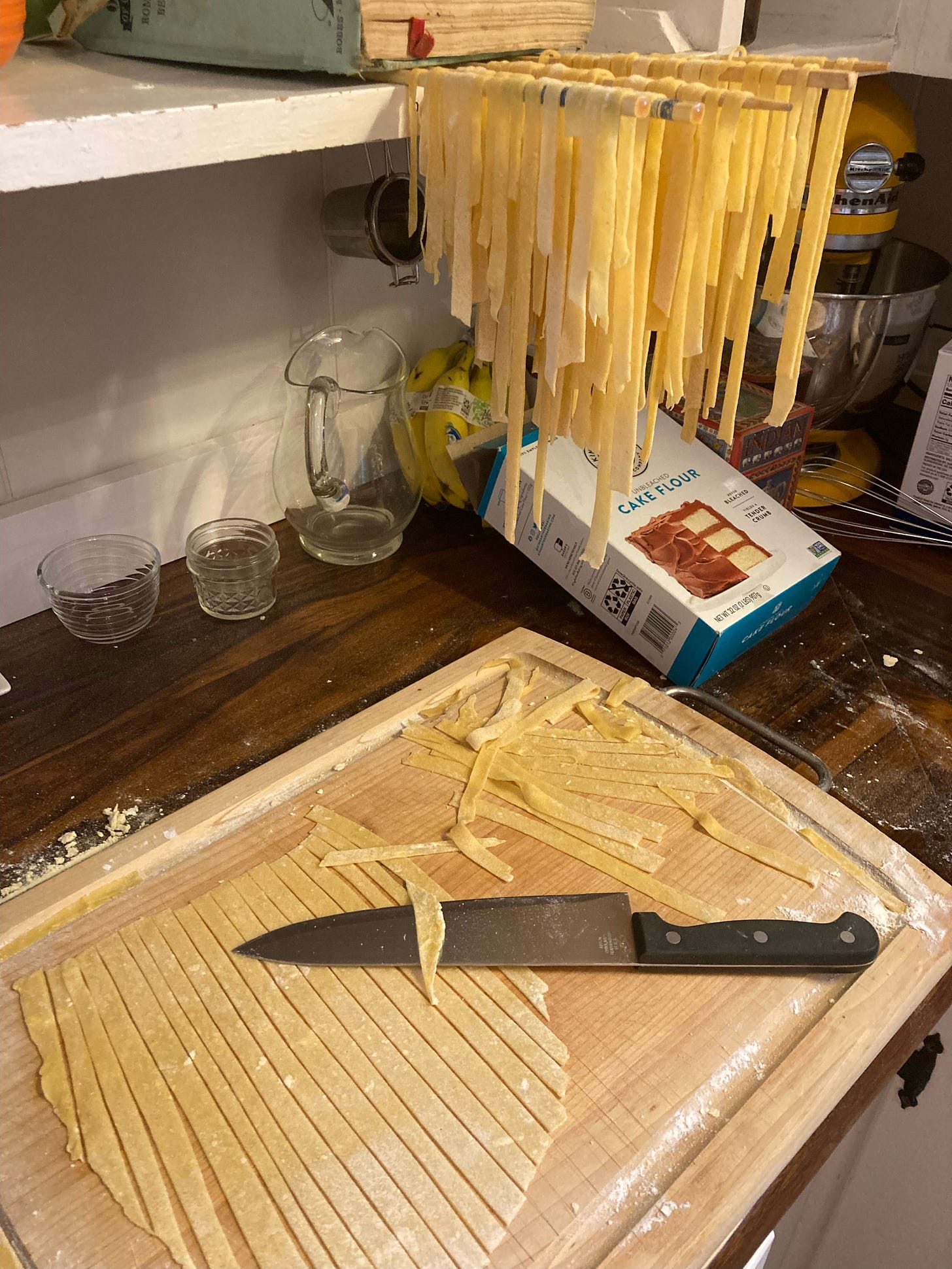 Freshly made pasta hangs from chopsticks while more is being cut on a wooden board on a counter, also visible is a box of Cake Flour from King Arthur Company.