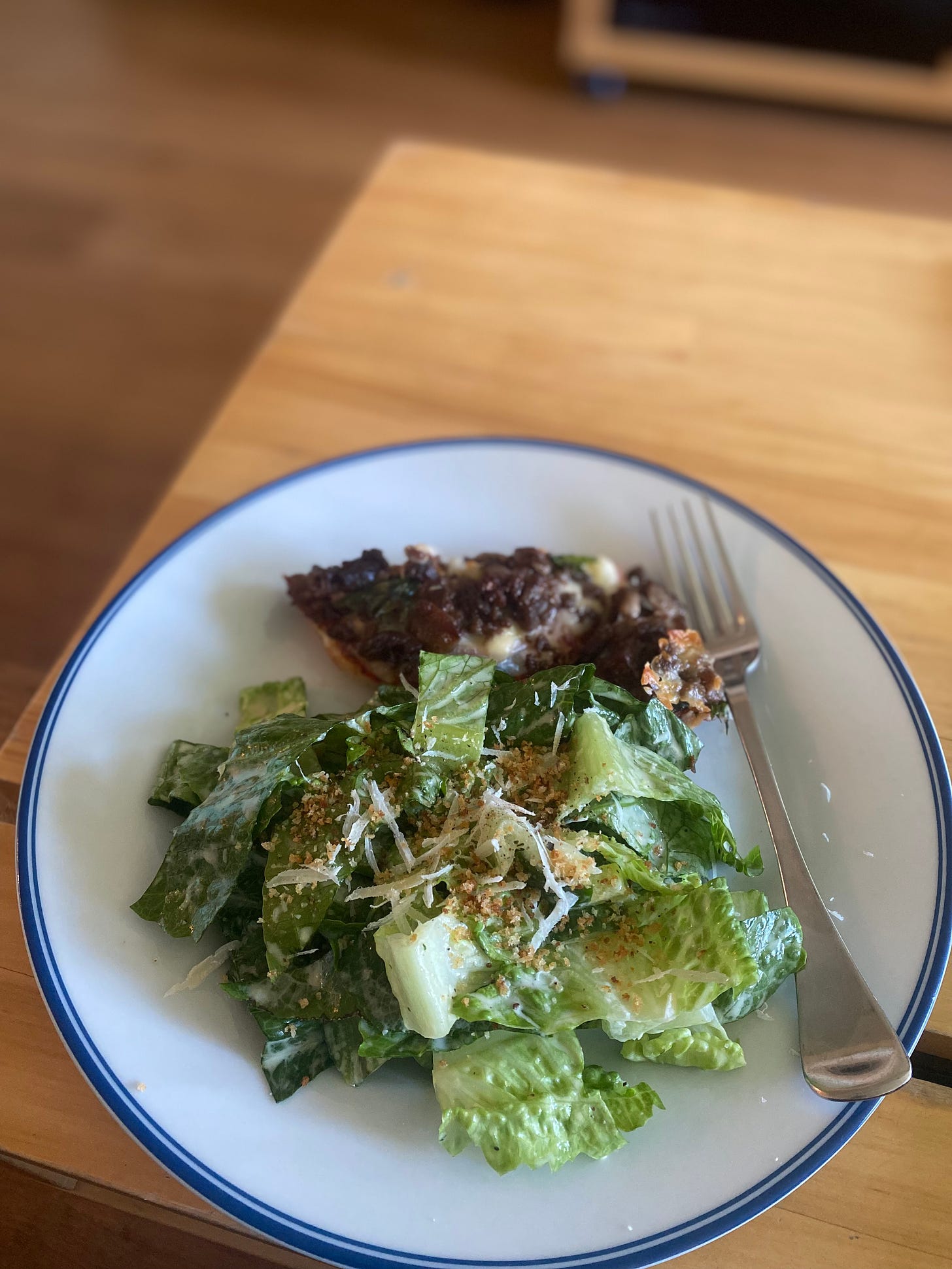 On a white plate with a blue rim, a romaine caesar salad topped with crispy breadcrumbs and grated parmesan. Behind it is half of a wagyu pizza bagel.