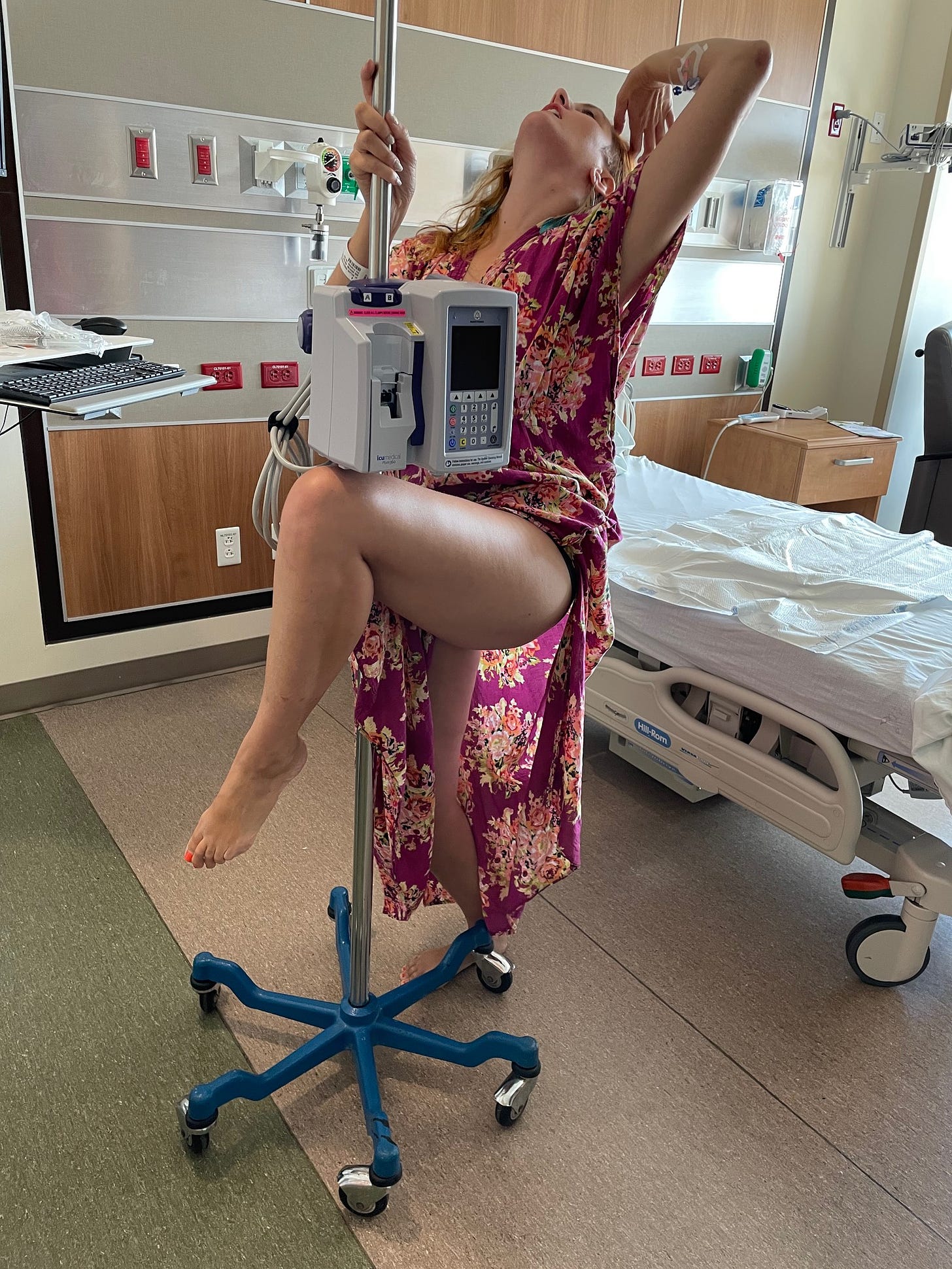 A thin white woman wraps her leg around an IV pole and arches her back toward a hospital bed behind her.