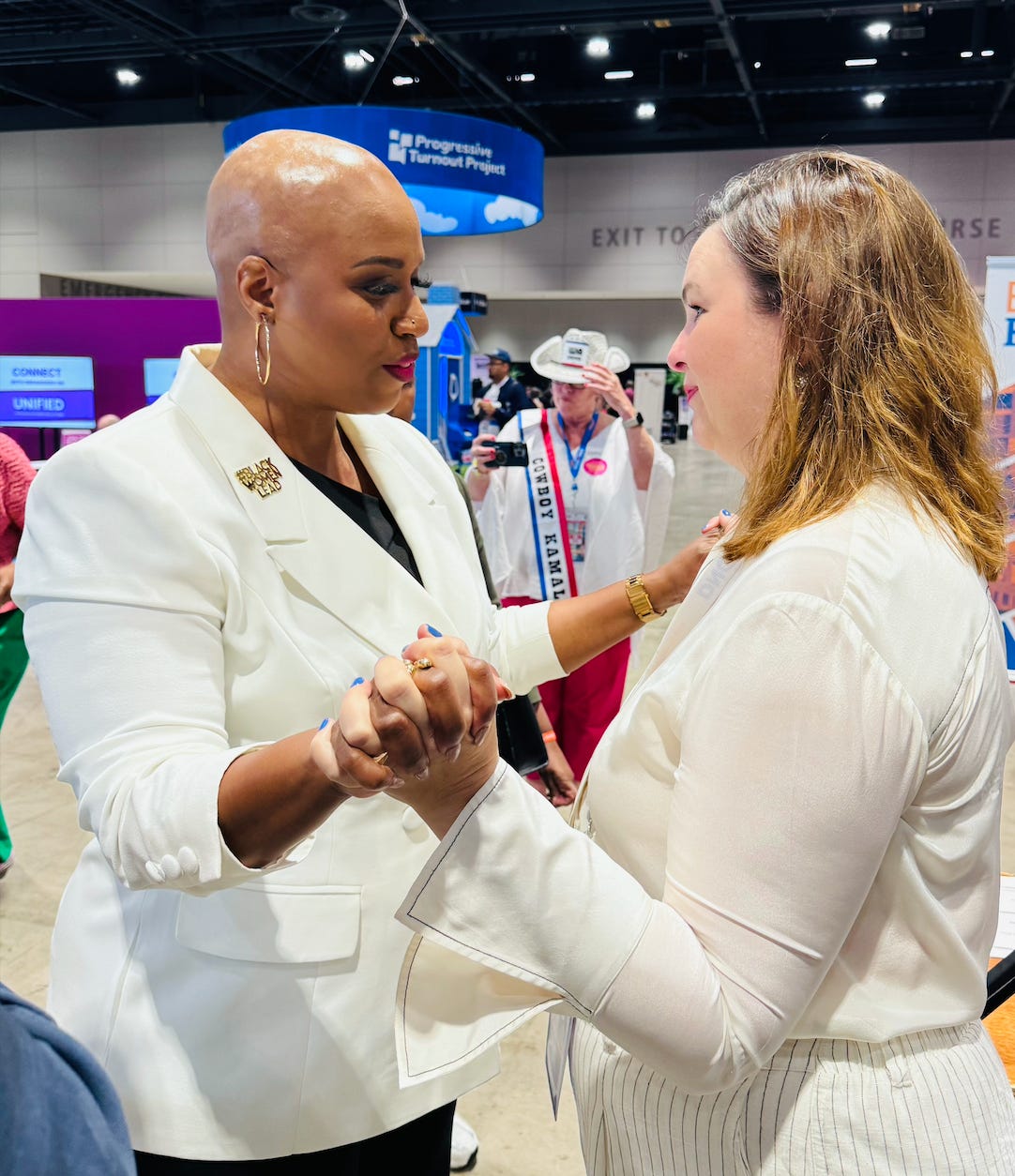 Congresswoman Ayanna Pressley and Amber Tamblyn stand facing each other, clasping each other's hands. Both wear white.