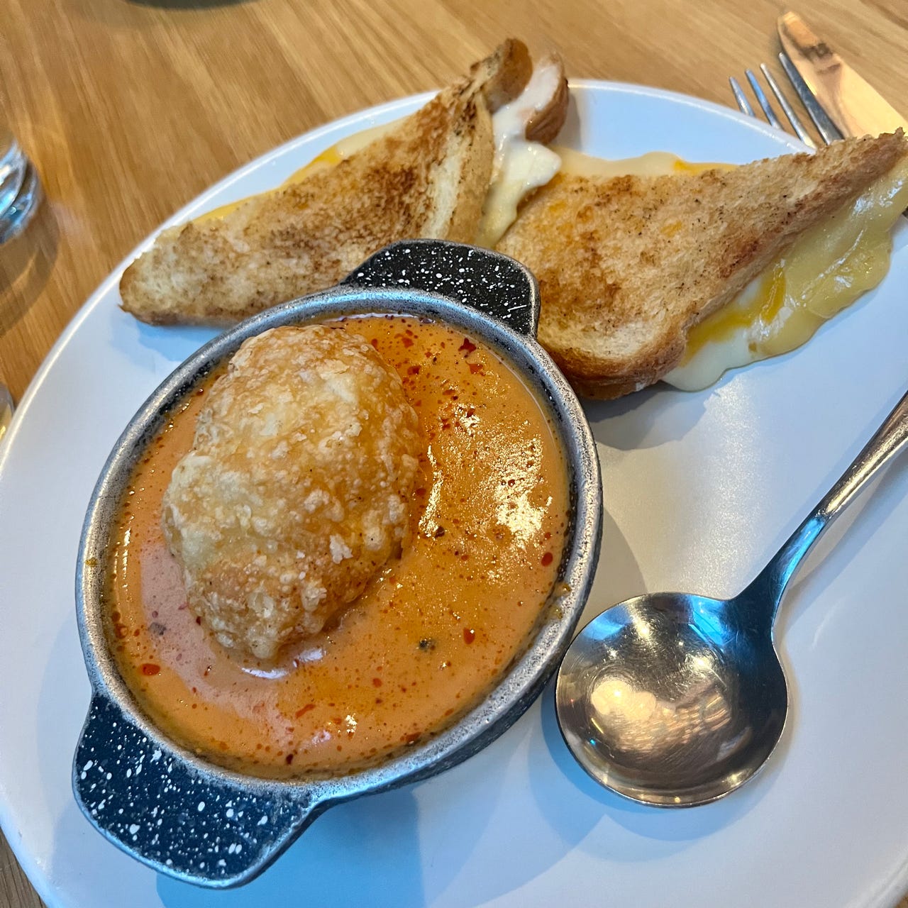 A bowl of orange tomato soup with a bready crouton in the middle. Next to it are two triangular slices of melty grilled cheese and a silver spoon.