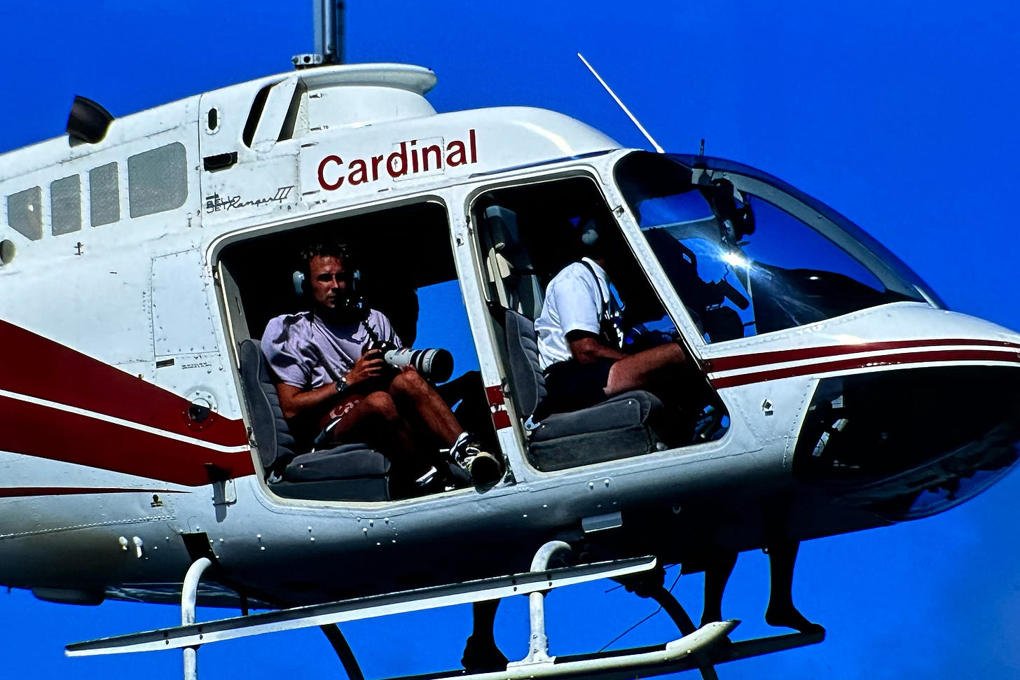 Photograph of a white helicopter with no doors in flight and a man sitting in the back holding a camera with a white lens.