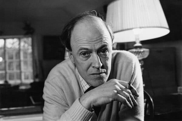 A black-and-white photograph of Roald Dahl, who is staring directly at the camera, with a cigarette in his right hand. A lamp is behind him. 