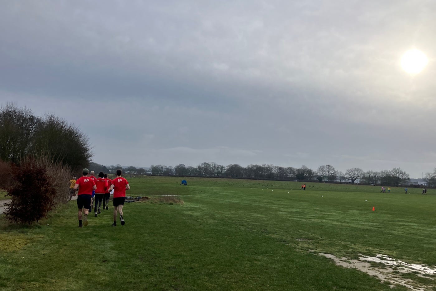 A wide shot of a field, runners in red ahead and the sun shining through clouds in the top right