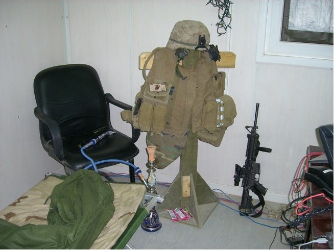 A color photo in the author’s office space in Camp Fallujah, Iraq in 2007. In the image is a military cot in the lower left, an office chair and a wooden rack holding the author’s combat equipment. On the rack is a helmet, ballistic vest with first aid kit and ammunition pouches attached. To the fight of the wooden rack is the author’s service rifle standing against the wall. To the right of the frame partially visible is the author’s desk with computer wires visible. 