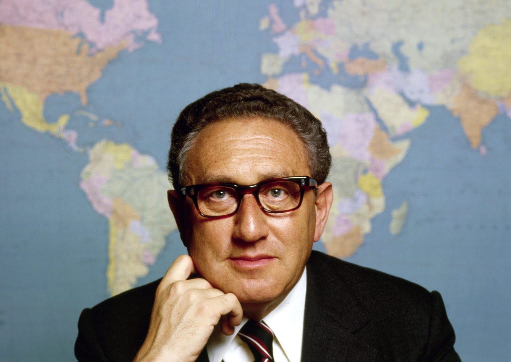 A color portrait of Ms. Kissinger in a dark suit jacket, white shirt and tie, his right hand pressed against his chin. A wall map of the world fills the space behind him. A color portrait of Ms. Kissinger in a dark suit jacket, white shirt and tie, his right hand pressed against his chin. A wall map of the world fills the space behind him.