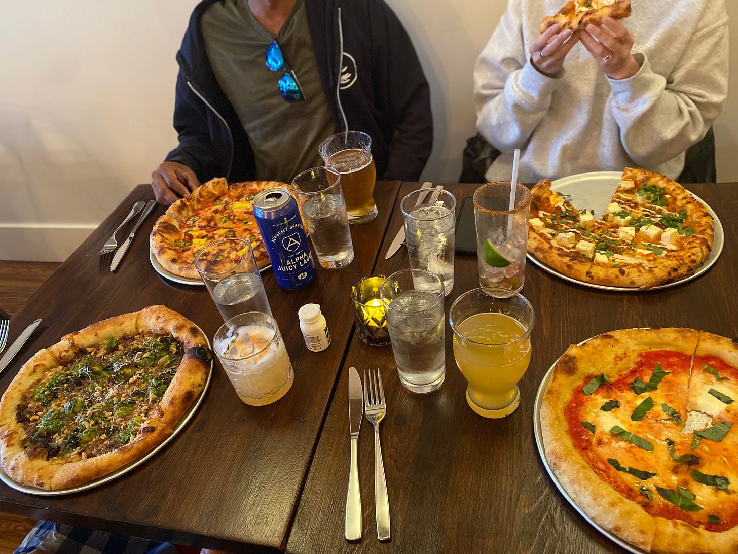 A table with four pizzas and a few drinks, as described below. Hiran and Tara (faces not visible) sit on the far side of the table.