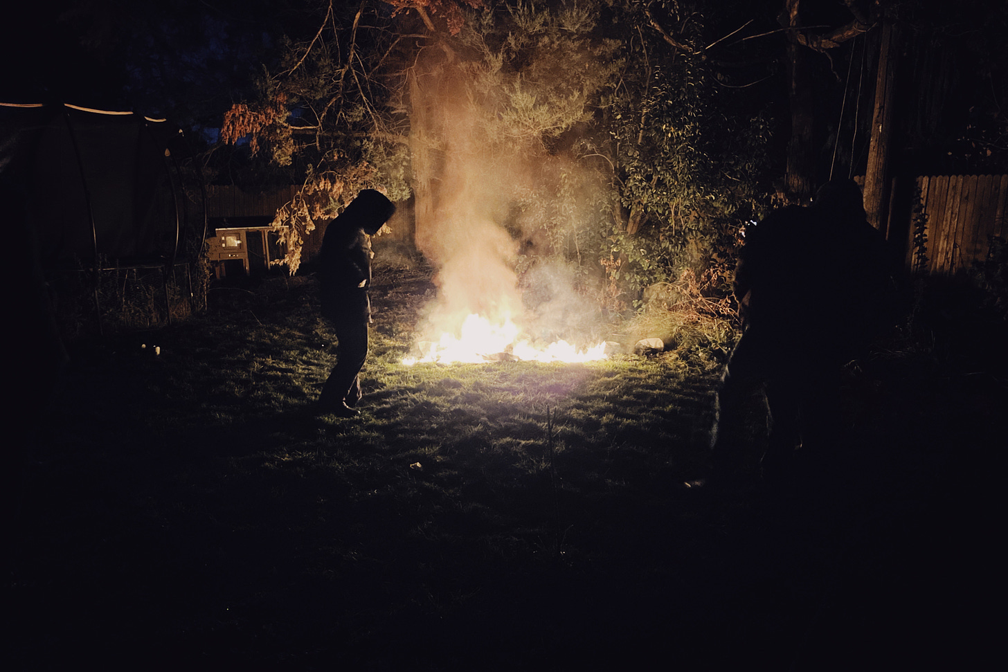 A photograph of the Patriot Front flag, which has been lit on fire and is burning on the ground. Almost nothing is left. Two shadowy figures stand off to the sides, illuminated by the fire. Their surroundings are dark.