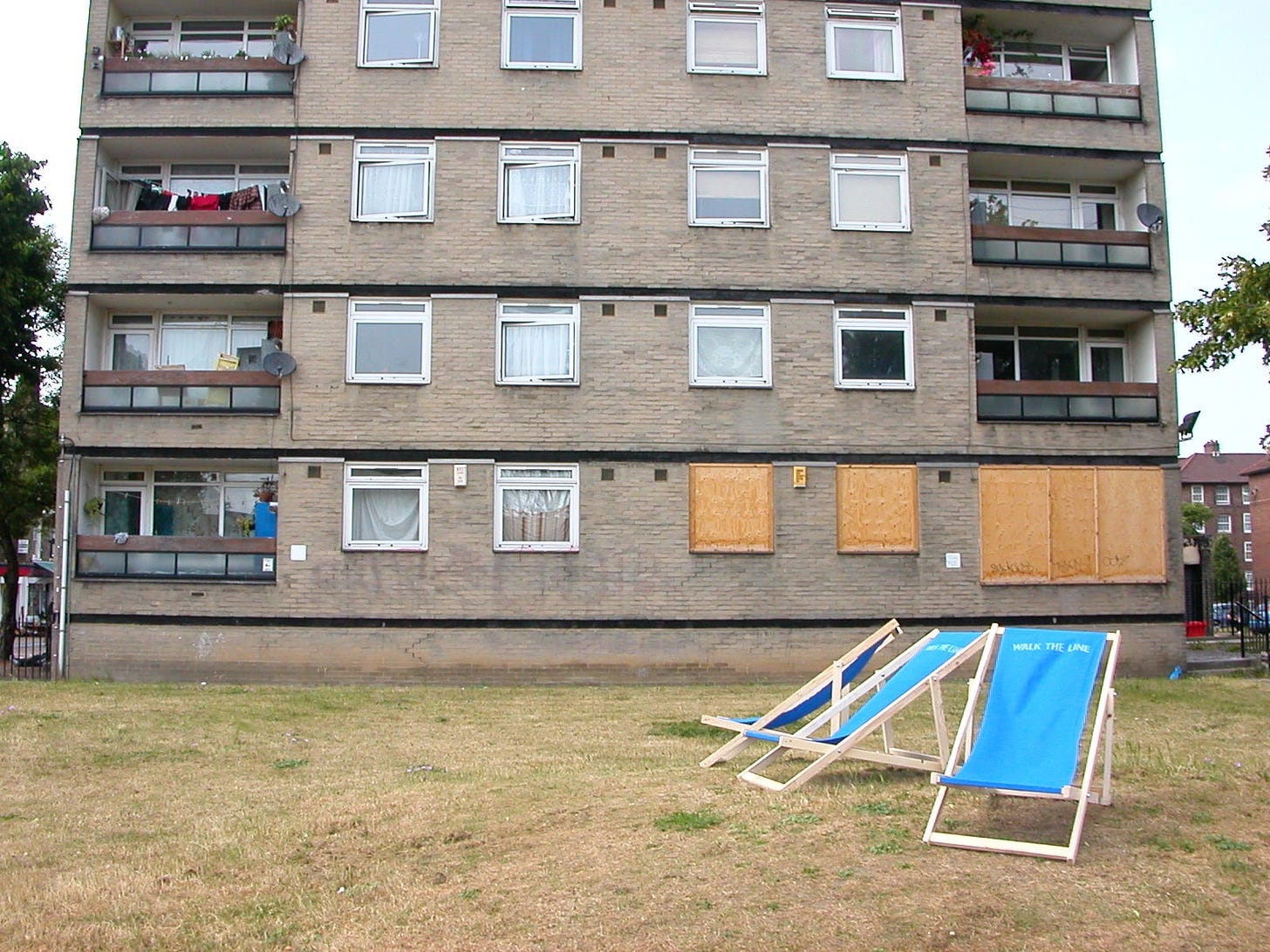 A tower block in East London, with one apartment's windows boarded up. Outside, on brown brittle grass, are three blue reclining cloth chairs with the words "Walk the Line".