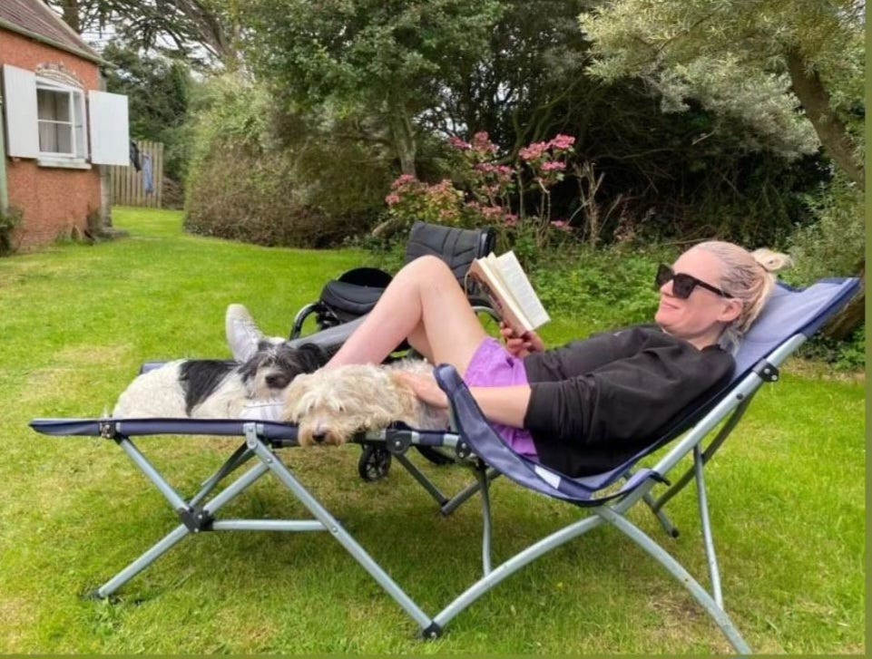 Wearing a pair of purple shorts, a black jumper and shades, I lie on a reclining chair with my two dogs lying under my legs. I'm reading a book and my wheelchair sits behind me in the garden. 
