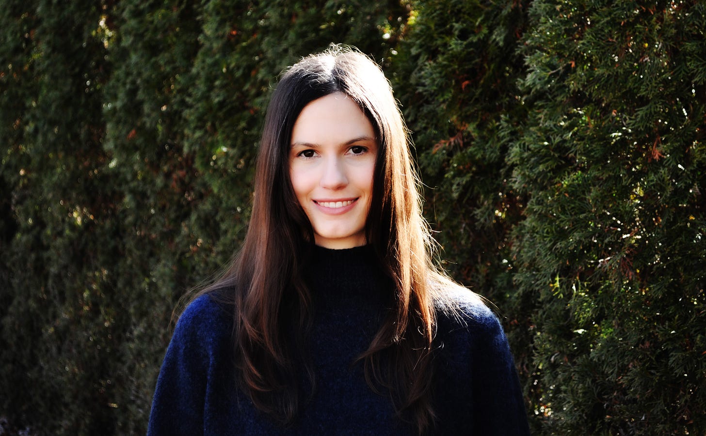 Headshot description: Olivia Muenz, a white woman with long brown hair and brown eyes, is smiling and wearing a navy sweater. She is in sunlight in front of rows of evergreen trees.