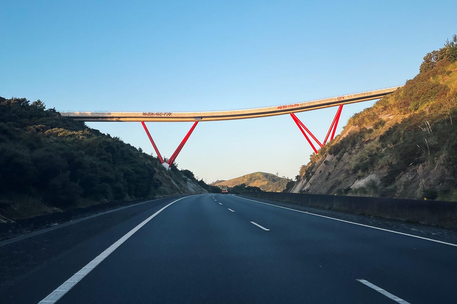 a late evening scene on a motorway, golden light hitting an unusual bridge with angled red struts, flanked by scrubby hills
