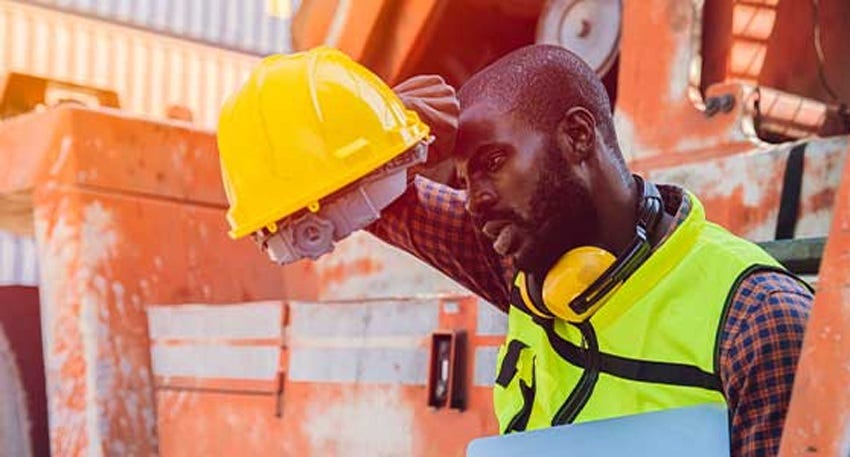 A Black man wearing a safety vest over a checked shirt leans against earthmoving equipment, removing his hard hat to wipe sweat off his forehead