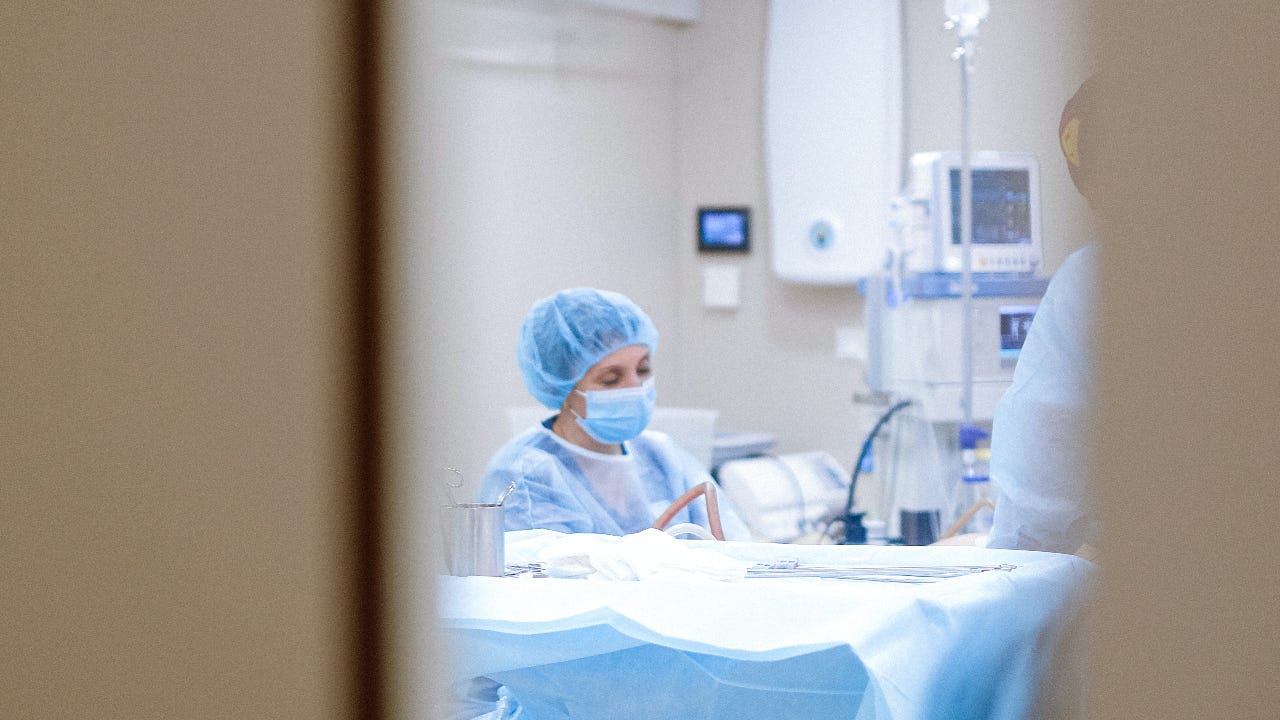 Someone in an operating room with a mask and hair net on the operating floor viewed through a door window.