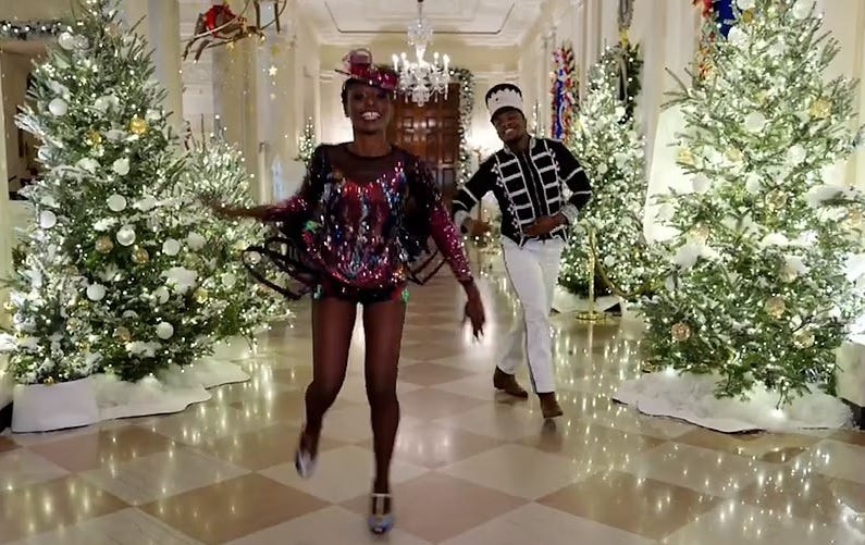 Video screenshot: two African American dancers, one costumed as a sequined sugar-plum fairy, the other as a toy soldier, tap[ dance in a tiled hall lined with sparkling Christmas trees