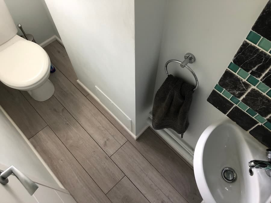 Small downstairs cloakroom bathroom toilet, sink, and towel rail, there is a green and black tile backsplash behind the sink. The towel rail is silver and has a dark grey towel hanging on it. 