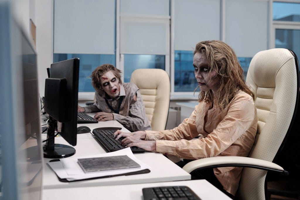 Young zombie businesswoman working at her desk, with a deceased male colleague in the background. Young zombie businesswoman working at her desk, with a deceased male colleague in the background.