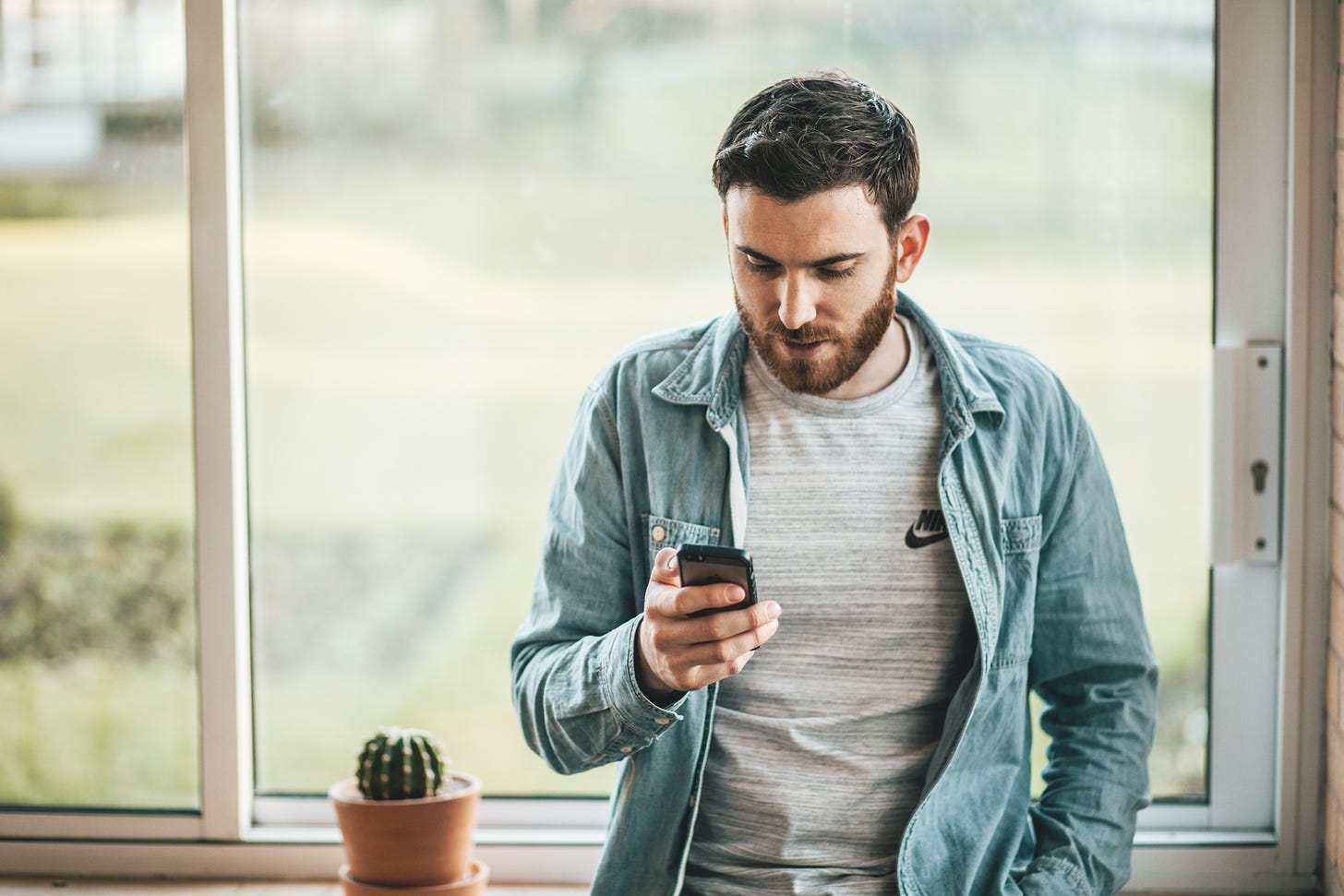 A man with an open blue denim shirt looks at his cell phone. He is standing in front of a window. 