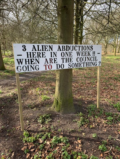 Caption of a sign nailed to two pieces of wood standing in front of a tree, sign saying, three alien abductions here in one week when are the council going to do something