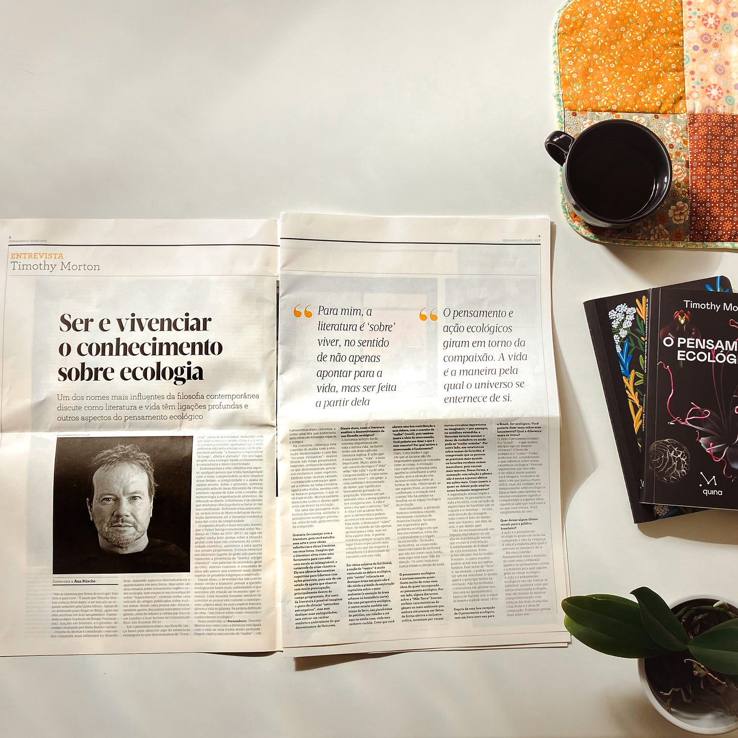 Um jornal aberto ao meio, com uma matéria sobre Timothy Morton, um livro da mesma autoria, um vaso de planta e uma xícara de café.