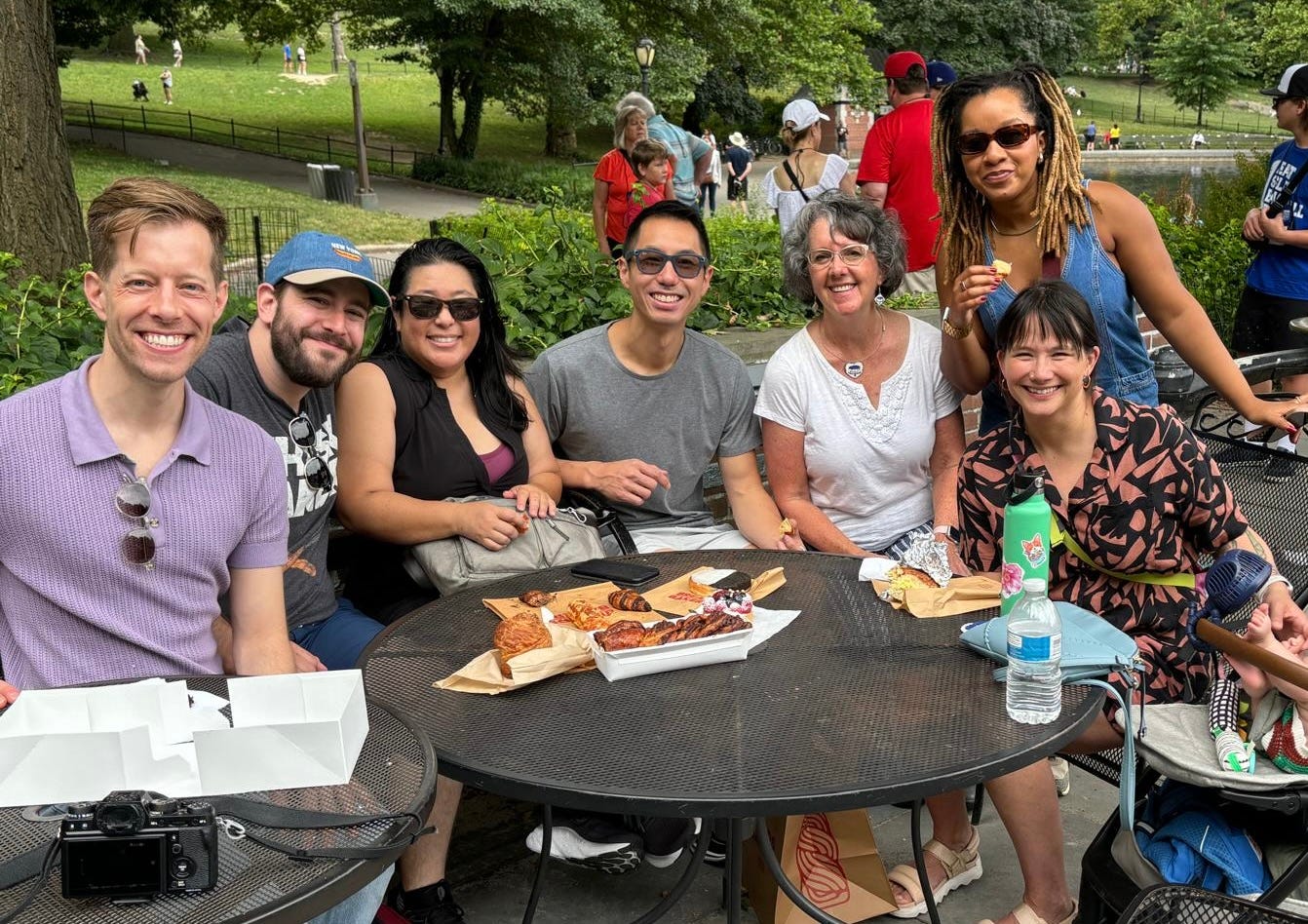 Most of the 2023 Great American Baking Show cast sitting at tables eating various pastries in Central Park.