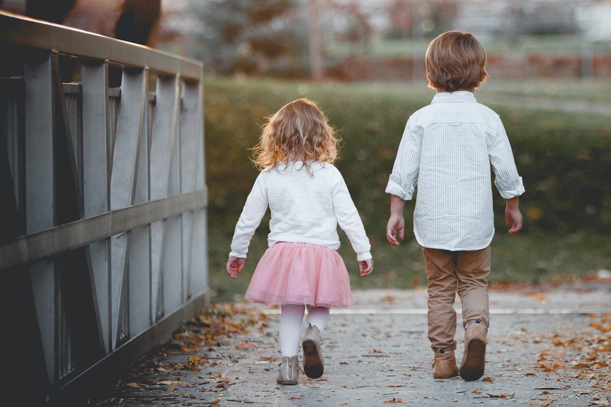 A boy and girl walk down a bridge, headed for a green grassy field.