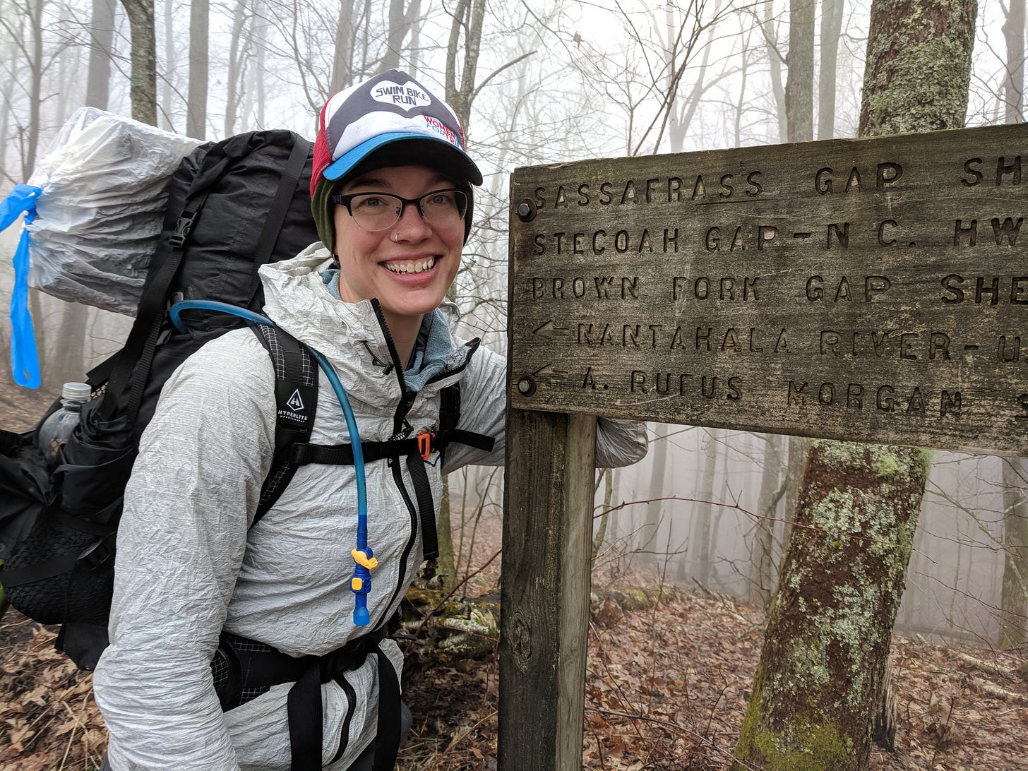 Another photograph of Kate from the Appalachian Trail. Here, she stands with one arm leaning against a sign for Sassafrass Gap in North Carolina. It's a foggy, wet day. The sleeping pad on her pack is covered in a trash bag, she wears a rain coat, but she still has a giant smile on her face.