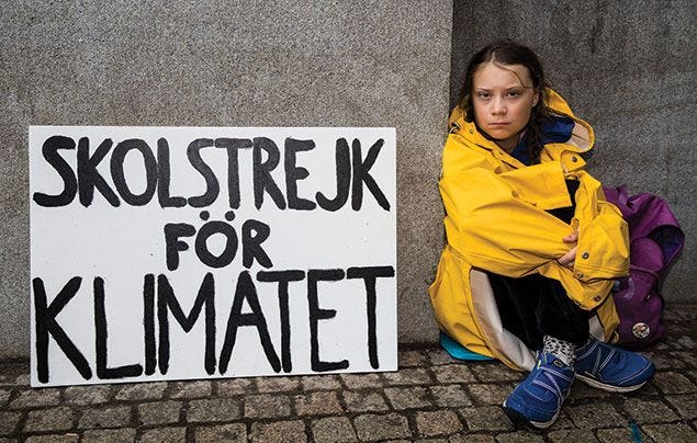 Greta Thunberg, a white teenager with blond braids, sits on the sidewalk against a concrete wall. She is wearing a bright yellow rain jacket and sitting next to a sign that says "Skolstrejk för Klimaetet"