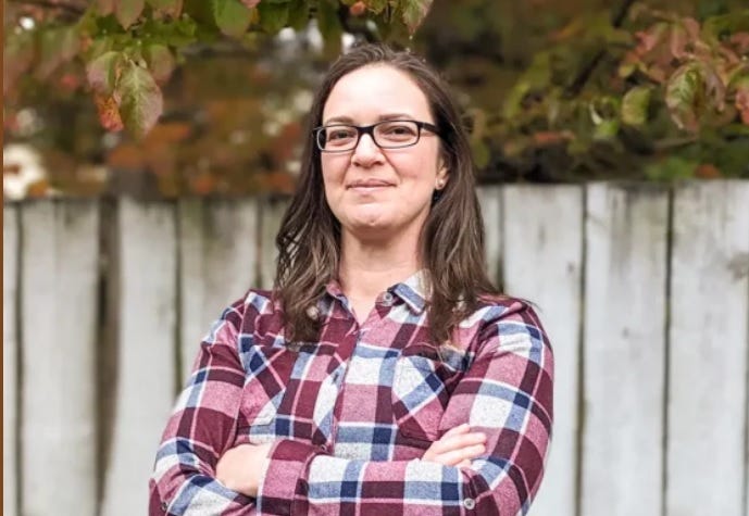 Sarah Taber, wearing glasses and a flannel shirt in muted red, white, and blue, stands with her arms crossed in front of a wooden wall. 
