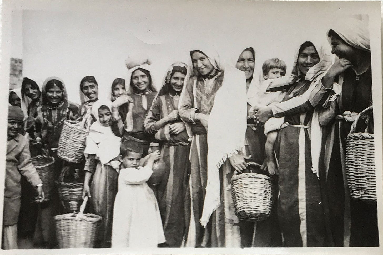 A sepia-toned old photo of a group of Palestinian women and children smiling at the camera.
