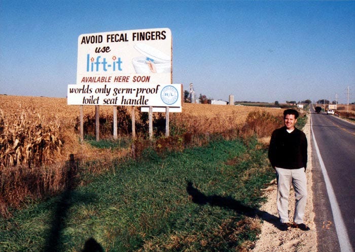 Image of a white man standing by the side of a highway next to a billboard that reads "AVOID FECAL FINGERS use Lift-it available here soon worlds only germ-proof toilet seat handle" 