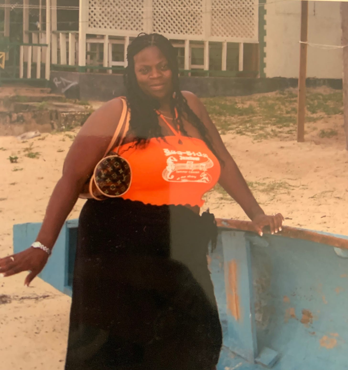 Black woman poses on a beach with a black skirt, orange halter top, leaning on a boat.