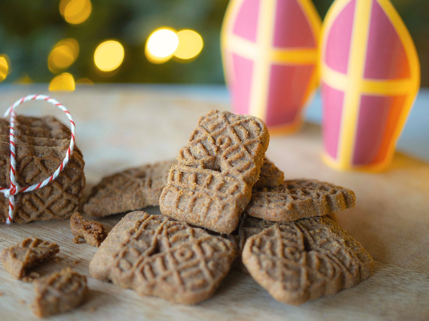 speculaas biscuits shaped lik windmills in a pile, some broken, a stack of them to the side, all on wooden board with St Nicholas hats in paper -kid's craft- in the background.