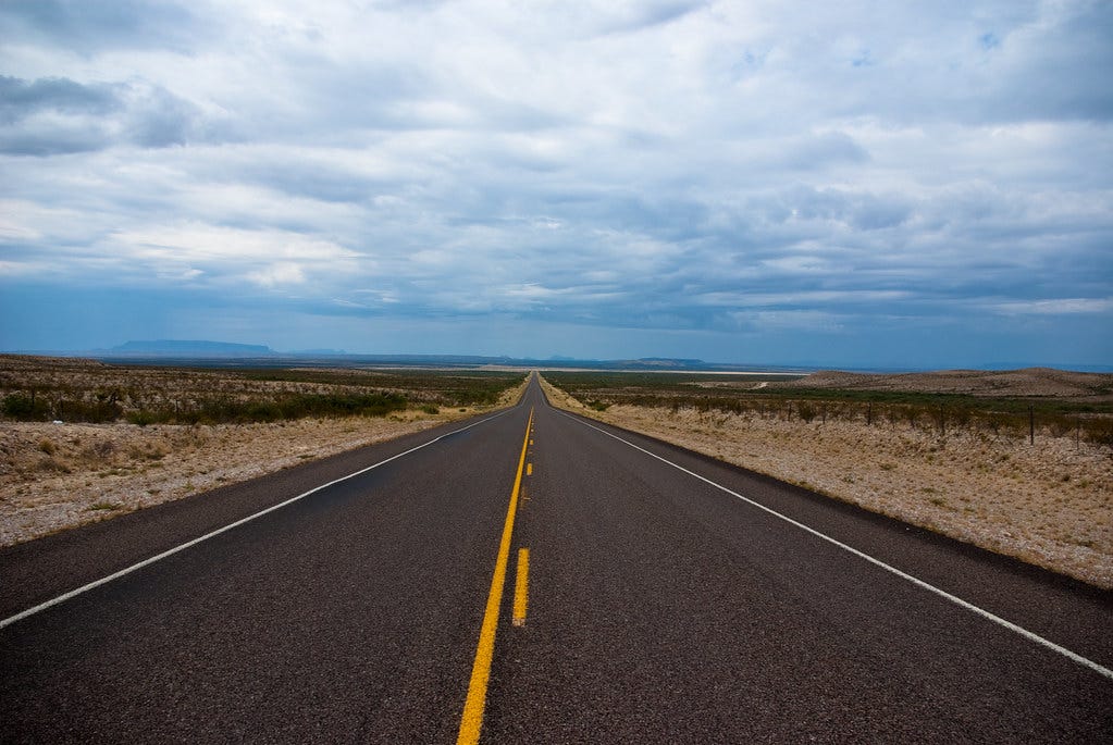 Two lane road disappearing into the horizon, through a dry Western landscape. "The Long Road" by Corey Leopold is licensed under CC BY 2.0. 