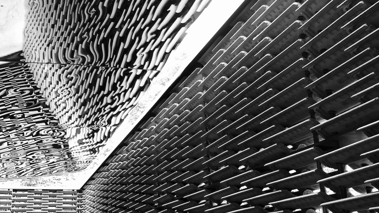 Black and white photograph of a modern architectural interior featuring horizontal slat-like wall panels and a mirrored ceiling. The slats create a repetitive, geometric pattern, which is reflected in the ceiling with a distorted, wavy appearance. A bright gap between the walls and ceiling reveals the sky outside. Black and white photograph of a modern architectural interior featuring horizontal slat-like wall panels and a mirrored ceiling. The slats create a repetitive, geometric pattern, which is reflected in the ceiling with a distorted, wavy appearance. A bright gap between the walls and ceiling reveals the sky outside.