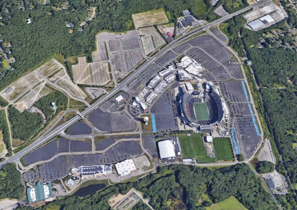 Satellite view of Gillette Stadium in Foxborough, Massachusetts, surrounded by vast parking lots and limited green spaces. The nearby Foxboro commuter rail station is visible to the right, illustrating the car-centric design and minimal transit accessibility of the area.