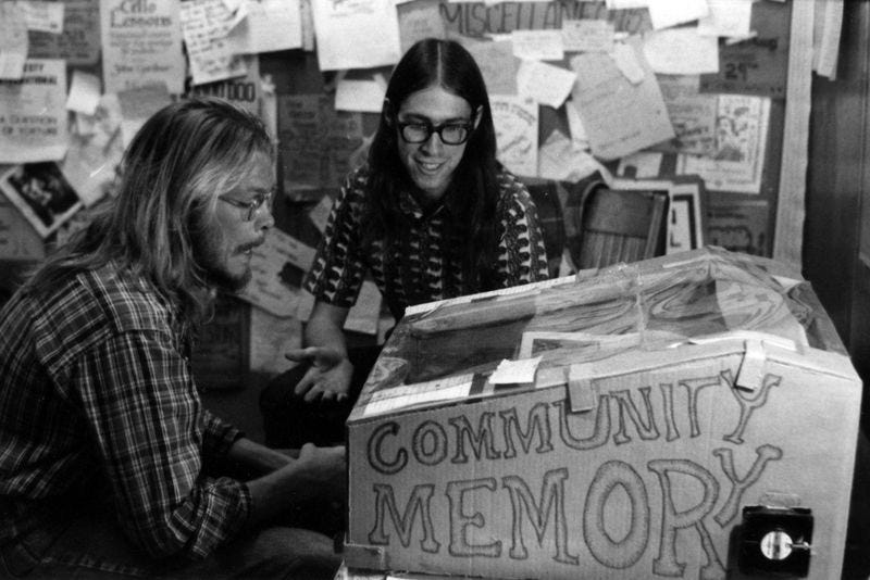 Two men with long hair sit in front of a cardboard box with "Community Memory" scrawled on the side