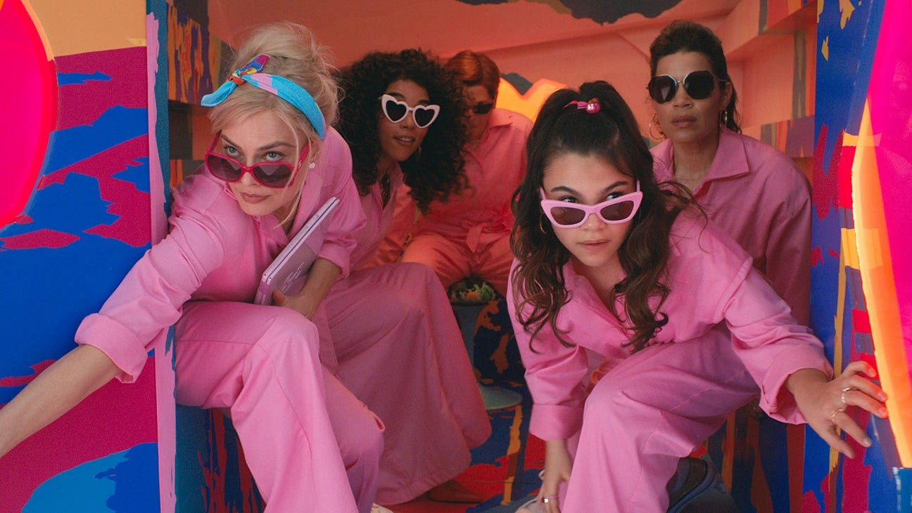 Out the back of a brightly colorful van, two women in pink jumpsuits and sunglasses are leaning out to exit. Behind them are two other women and a man also clad in pink jumpsuits and sunglasses.
