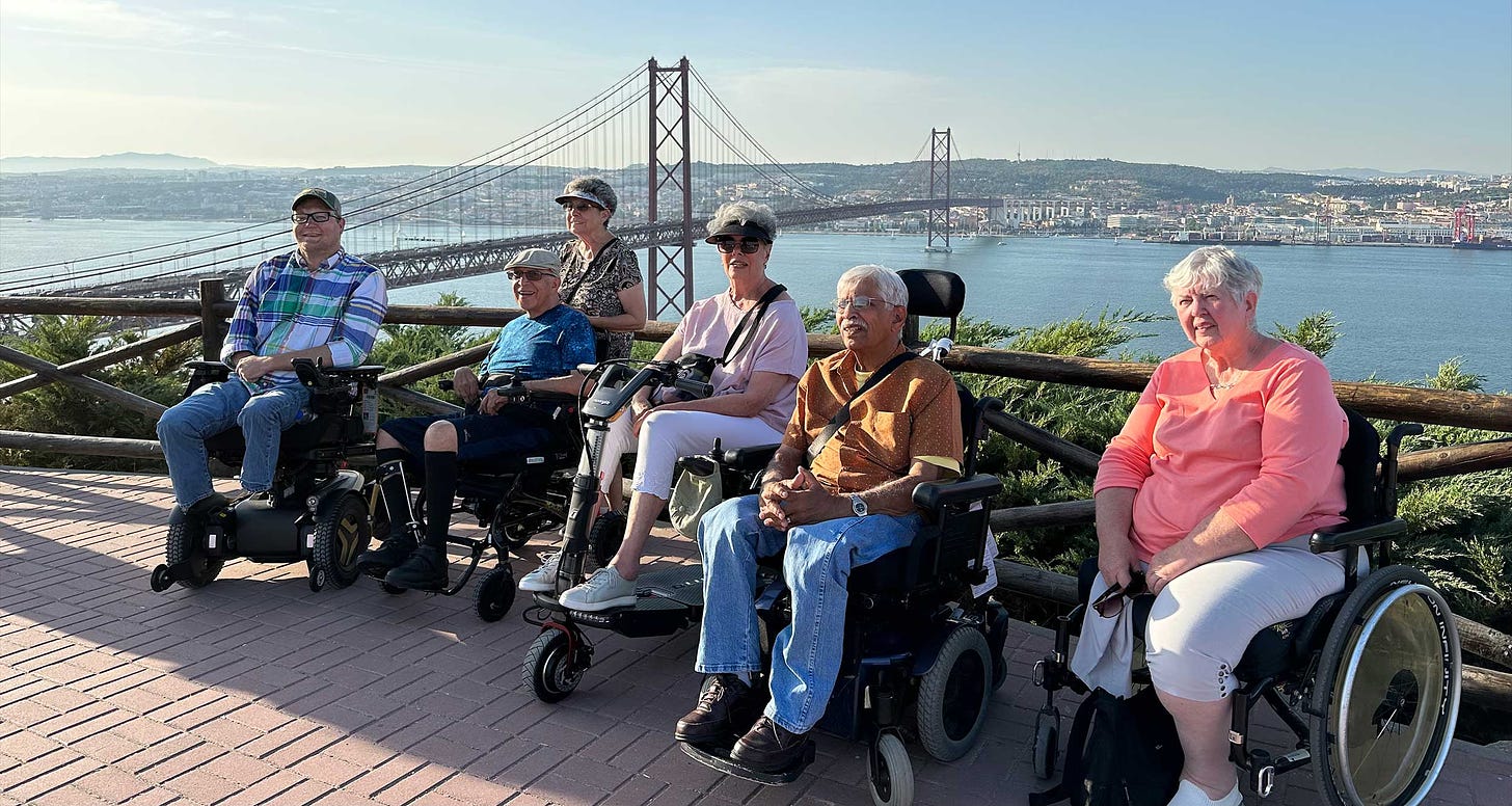 Group of wheelchair users and disabled travelers pictured in front of a bridge in Portugal, with the Lisbon skyline in the background.