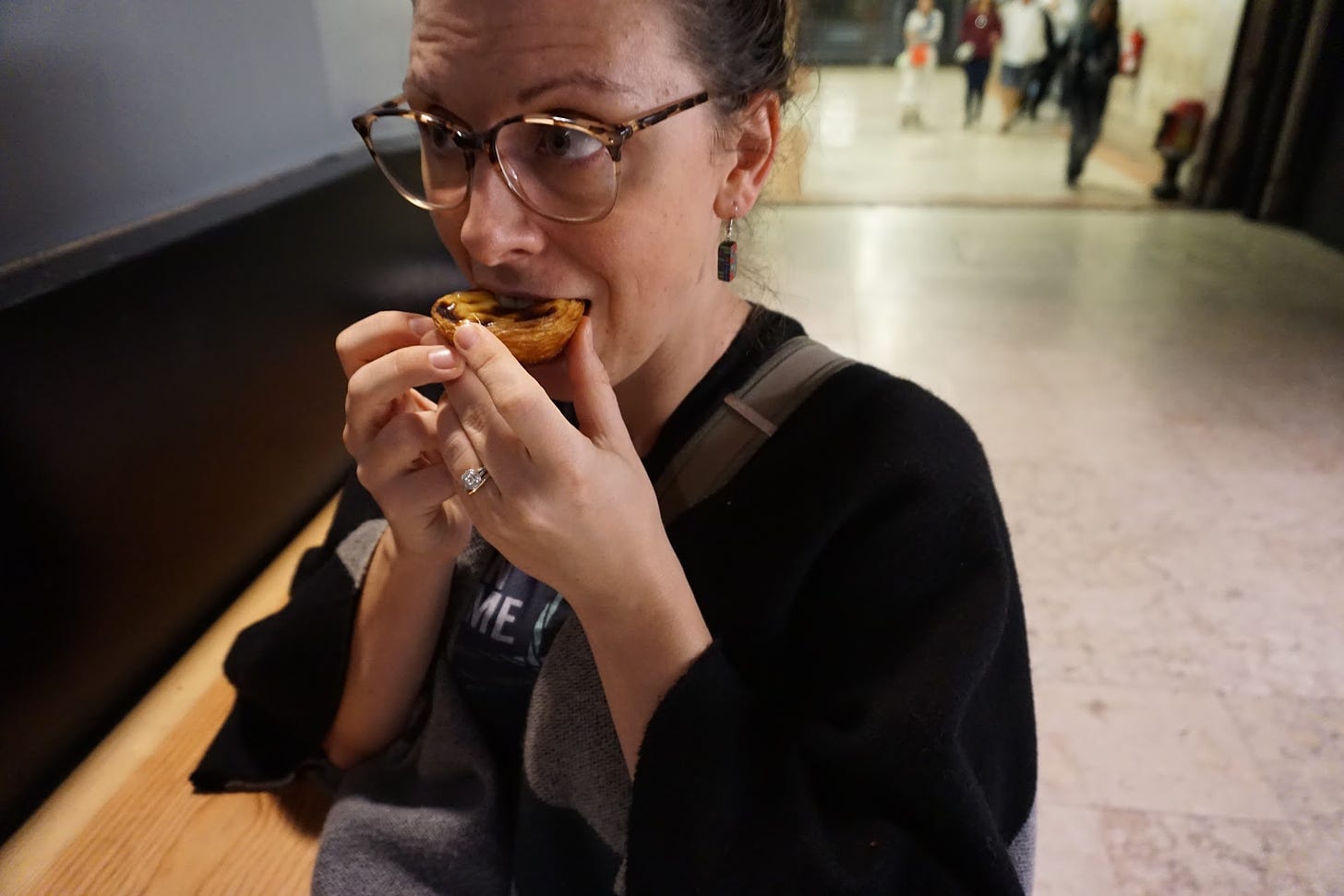 A woman (me) in a shawl looking away from the camera and eating a pastel de nata.