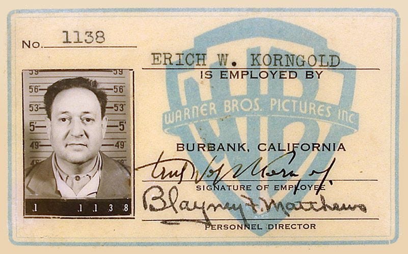 Photo of a 1940s-era employee ID featuring a black and white photo of a man wearing a suit, alongside text saying "Erich W. Korngold is employed by Warner Bros. Pictures Inc. / Burbank, California and two signatures Photo of a 1940s-era employee ID featuring a black and white photo of a man wearing a suit, alongside text saying "Erich W. Korngold is employed by Warner Bros. Pictures Inc. / Burbank, California and two signatures