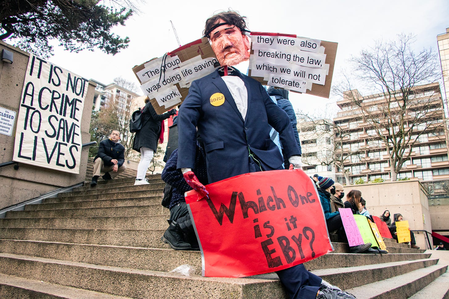 A papier mache David Eby sits on some steps outside at a protest. Over one shoulder, a sign says "The group was doing life-saving work." A sign to his right, says "they were also breaking the law, which we will not tolerate." Behind and to his left a group of protestors sit on the steps holding signs. To his right, a sign says "It is not a crime to save lives."